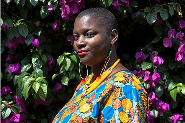 A woman in a colorful patterned top, bright jewelry and makeup, and a shaved head stands in front of a wall of flowering vines.
