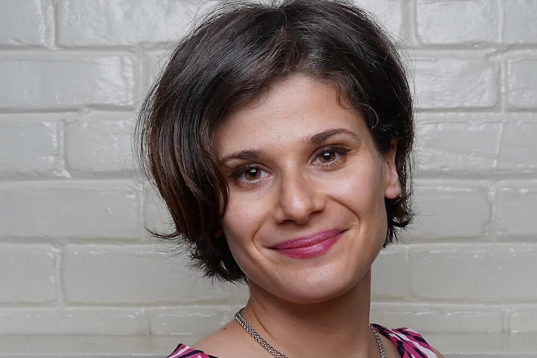 A woman with cropped brown hair, a patterned pink tank top, pink lipstick, and a silver chain poses in front of a white brick wall.