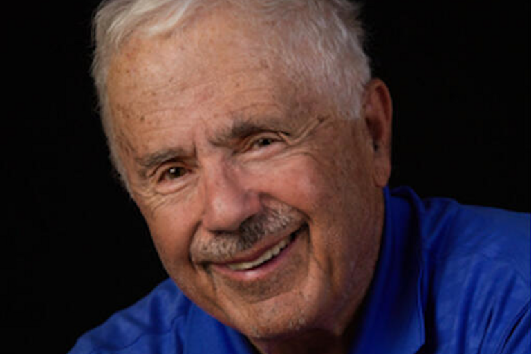 Close-up portrait of an elderly man smiling, wearing a blue polo shirt against a black background.