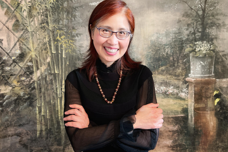 A smiling woman with red hair, wearing glasses and a black top, stands in front of a painted garden backdrop with bamboo plants and a stone planter.