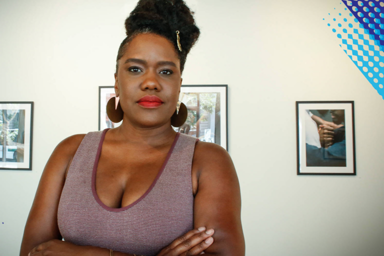 Person with arms crossed, wearing a light purple top and large circular earrings, standing in front of a grey wall with framed photographs.
