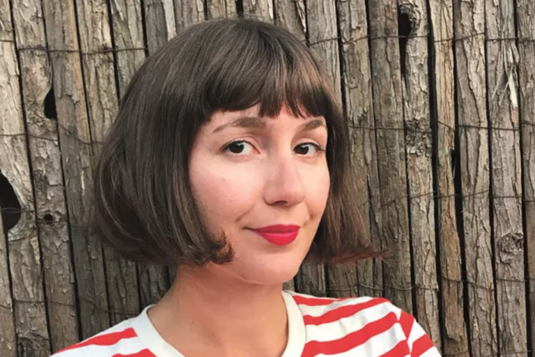 Portrait of a person with chin-length brown hair and red lipstick in front of a background of weathered wooden planks.