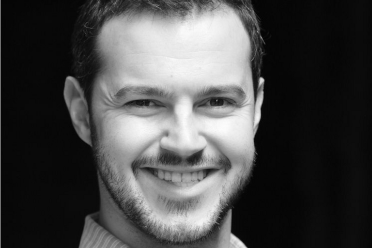 Black and white portrait of a smiling man with short, dark hair and a beard, wearing a striped dress shirt.