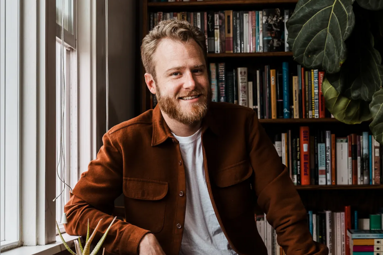 Man with short, wavy hair and a beard, wearing a brown jacket over a white T-shirt, standing next to a window and a bookshelf filled with books.