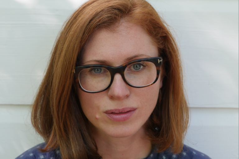 Close-up portrait of a person with red hair and dark-rimmed glasses against a white background.