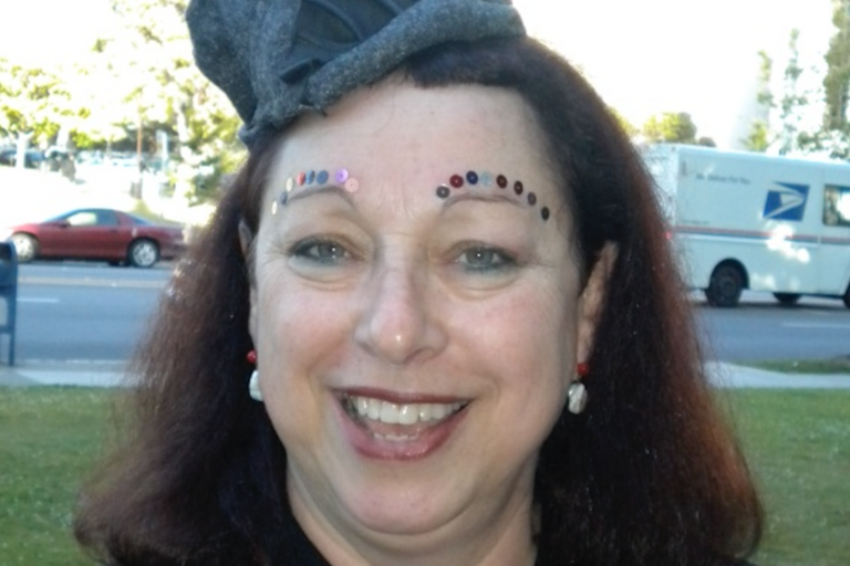 Close-up of a smiling person with decorative sequins above their eyebrows and a small grey hat on their head.