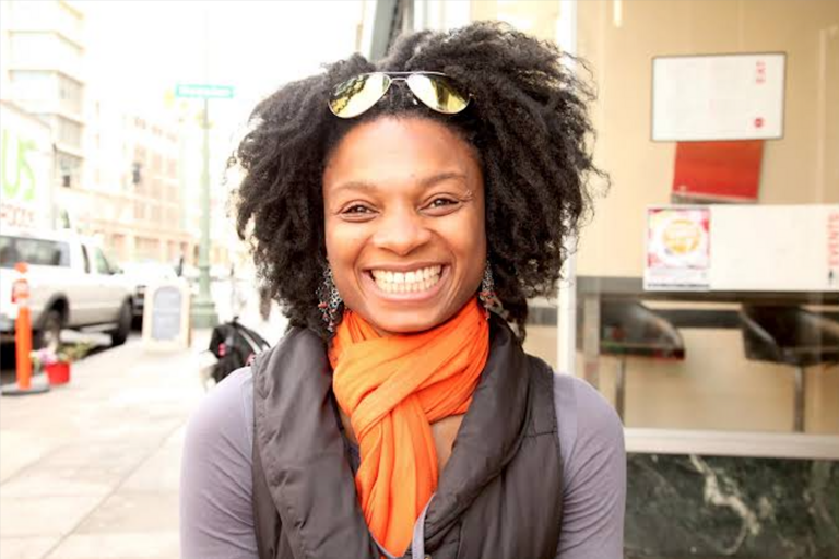 Smiling individual with curly hair wearing an orange scarf on a city sidewalk.
