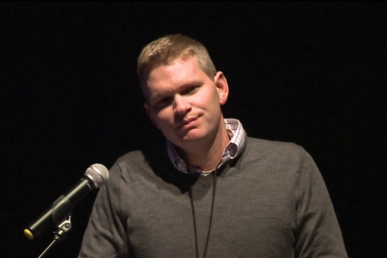 Man in a gray sweater speaking into a microphone against a dark background.