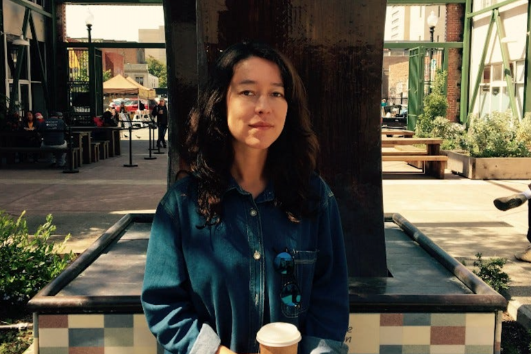 Person with long dark hair in a blue denim shirt holding a coffee cup in an outdoor industrial-style setting.