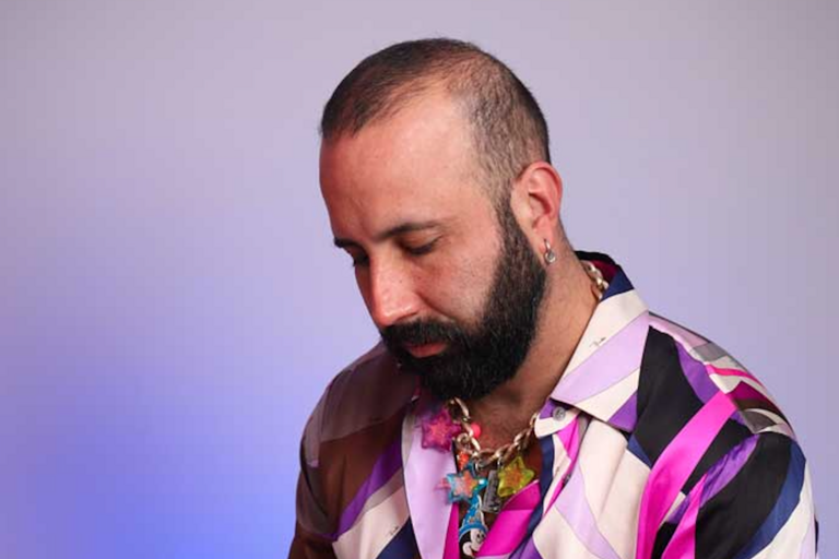 Person with a shaved head and beard, wearing a colorful striped shirt and a chunky necklace with star-shaped ornaments, looking downward against a blue and violet gradient background.