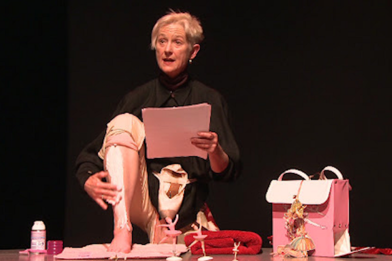 An older adult sits on a stage applying a white substance to their bandaged leg, with various items like a pink bottle and handbag in front of them.