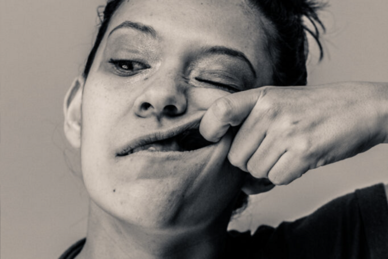 Black and white image of a woman with a bun in a T-shirt, her left pointer finger hooking the inside of her cheek.