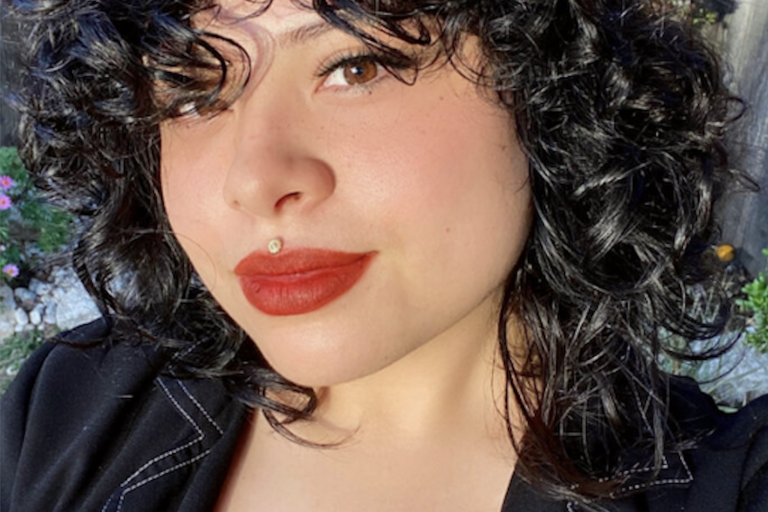 A person with curly hair and a red lip wears a black top, smiles towards the camera