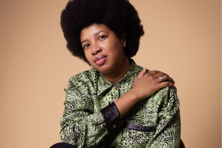 Person with afro sitting on chair in a green button-up, arms crossed over torso.