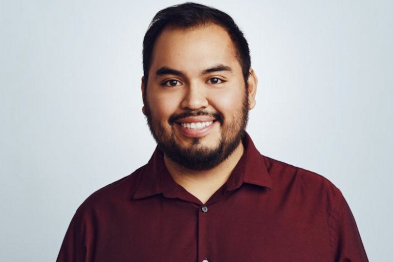 Smiling person in a maroon button-up shirt against a light blue background.