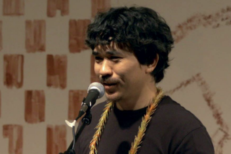 Man with a braided necklace speaking into a microphone against a patterned background.