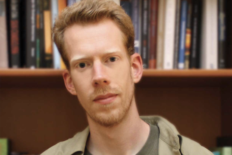 Young man with short hair and a beard in front of a bookshelf.