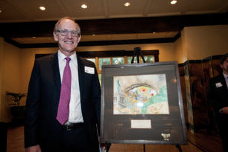 A man in a suit stands next to an easel displaying a colorful framed artwork in a well-lit room.