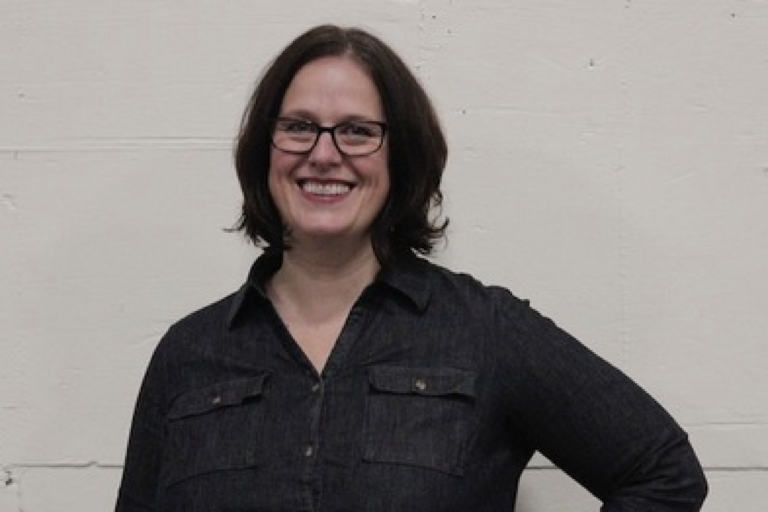Person with glasses and mid-length brown hair smiling, wearing a dark long-sleeved shirt, standing against a light-colored wall.