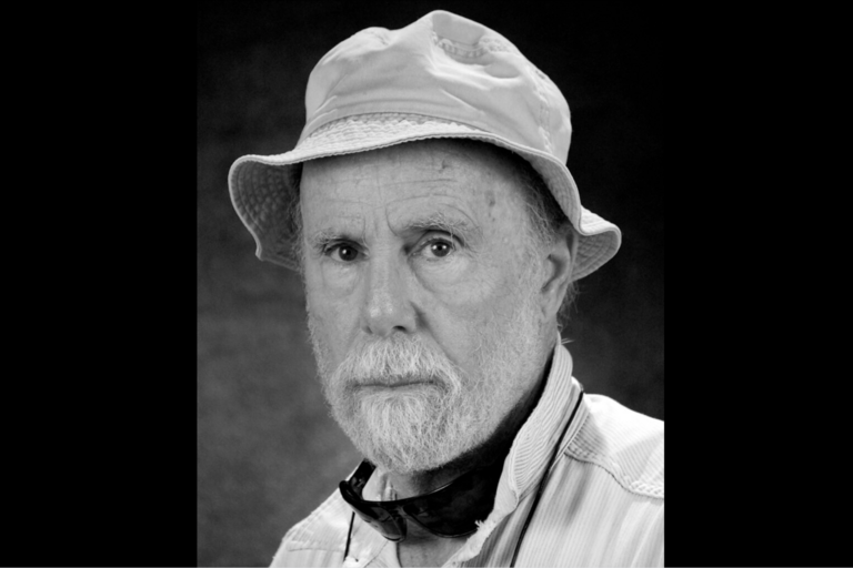 Black-and-white portrait of an older man wearing a bucket hat and a light-colored, striped shirt, with sunglasses around his neck.