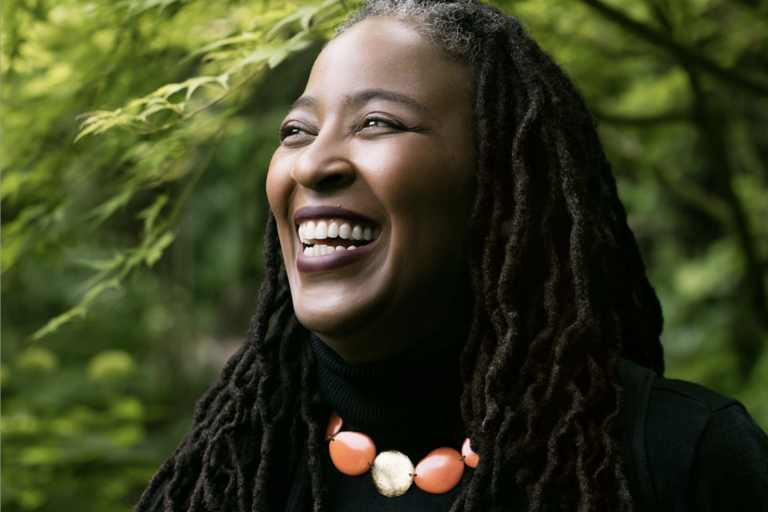Close-up of a smiling person with long hair, wearing a black turtleneck and an orange and gold beaded necklace, set against a greenery background.