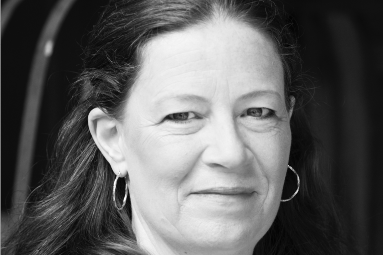 Black and white close-up portrait of a middle-aged individual with long hair and hoop earrings, looking at the camera with a subtle smile.