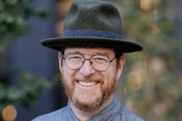 Close-up of a smiling person wearing a green fedora and round glasses.