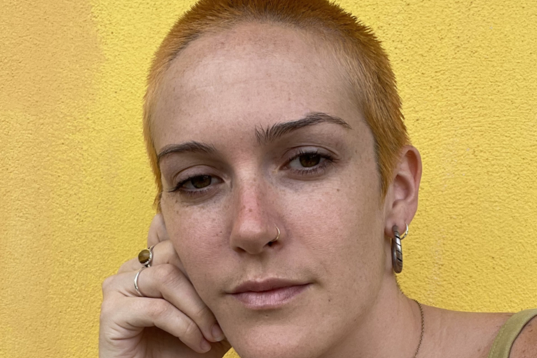 Close-up of a person with short orange hair, nose ring, and hoop earrings against a yellow wall.