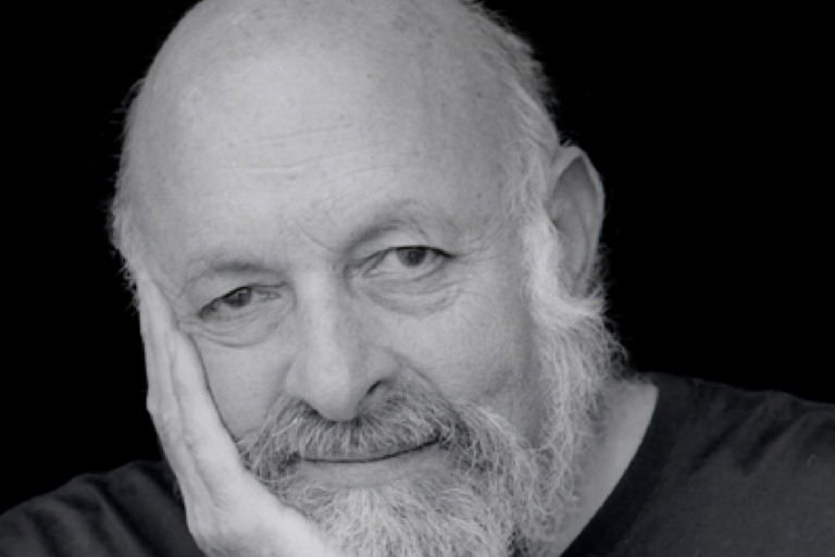 Close-up black-and-white photo of an older man with a bald head and white beard, resting his head on his left hand, against a black background.