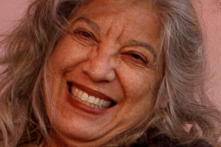 Close-up of an older person with long, wavy gray hair, smiling broadly with a pink background.