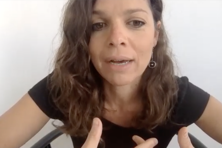 Woman with curly brown hair and black top speaking and gesturing with hands against a white background.