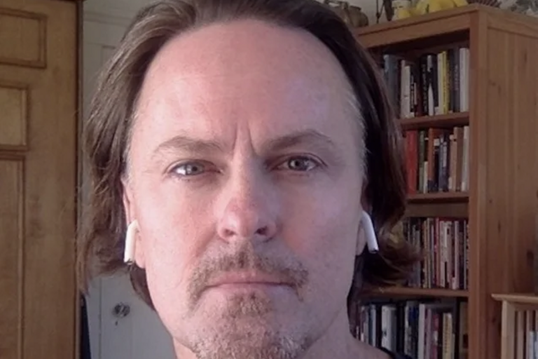 Close-up of a man with light brown hair and a short goatee, wearing white wireless earphones, with a bookcase in the background.