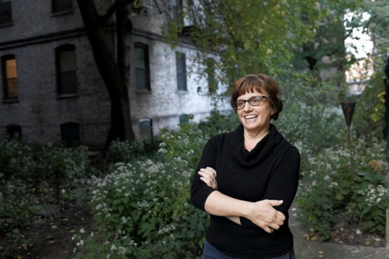 Middle-aged woman with glasses standing in front of a grey brick building, surrounded by a garden with green plants and white flowers.