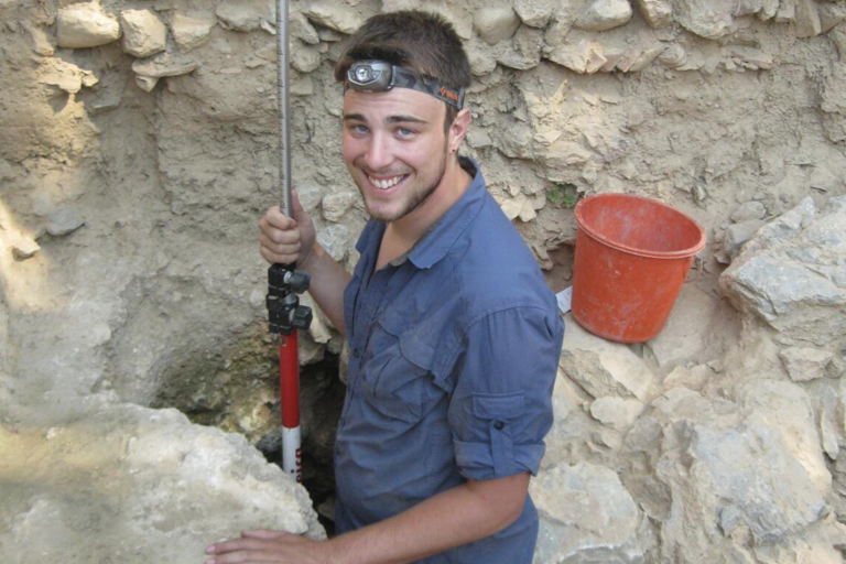 Person wearing a blue shirt and headlamp, standing in a dig site and holding a leveling rod, with an orange bucket nearby.