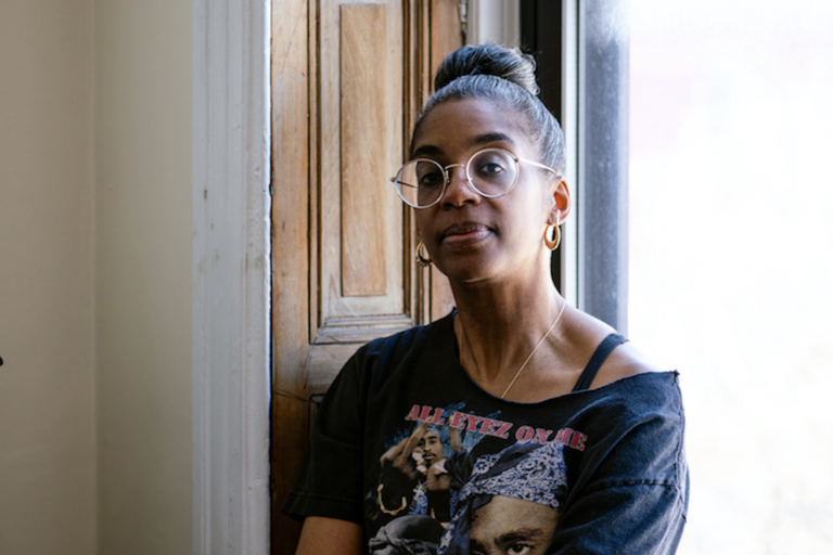 Woman wearing glasses and a black T-shirt with "ALL EYEZ ON ME" text, seated next to a window and wooden door.