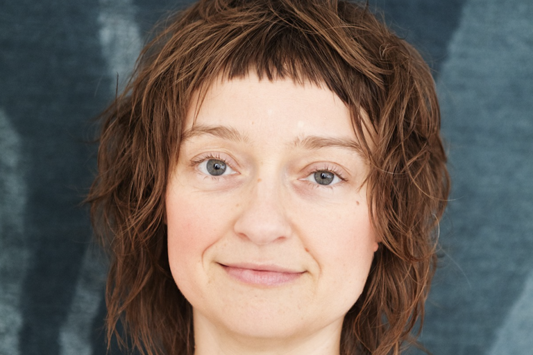 Close-up portrait of a person with light blue eyes, light brown wavy hair, and a neutral expression against a textured blue background.