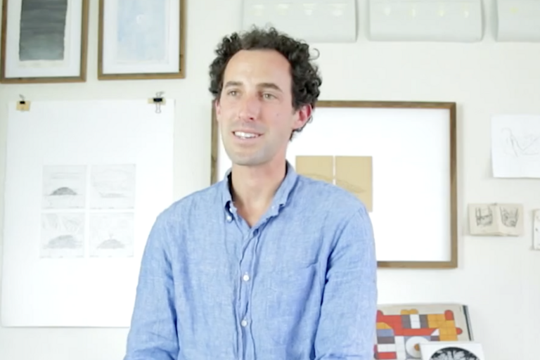 Person in a blue shirt sitting in front of a wall with framed pictures and drawings.
