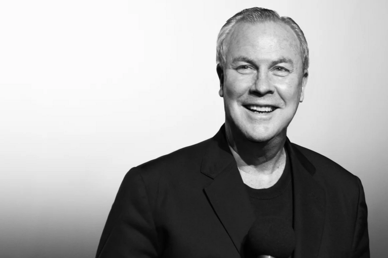 Black-and-white portrait of a smiling man in a dark blazer, holding a microphone.