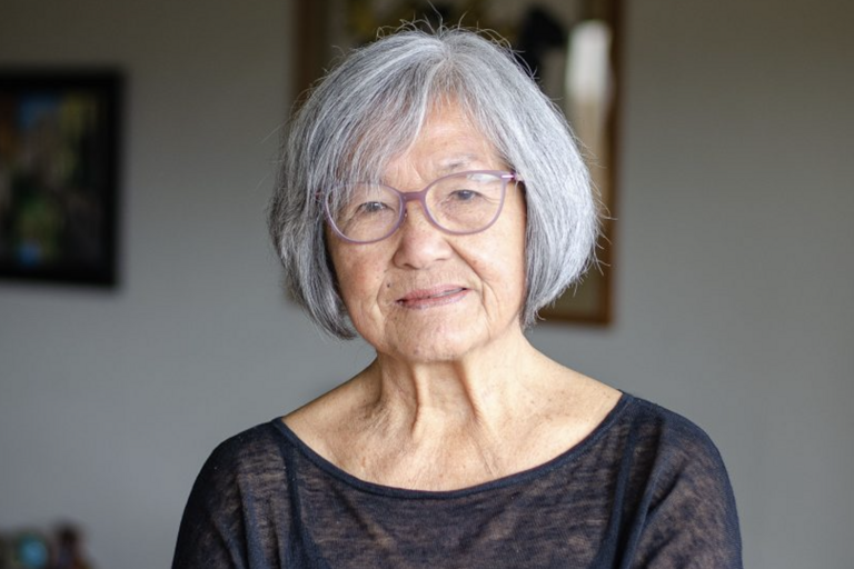Elderly woman with gray hair and purple-framed glasses, wearing a black semi-sheer top. Indoor setting with blurred art pieces on the wall behind her.