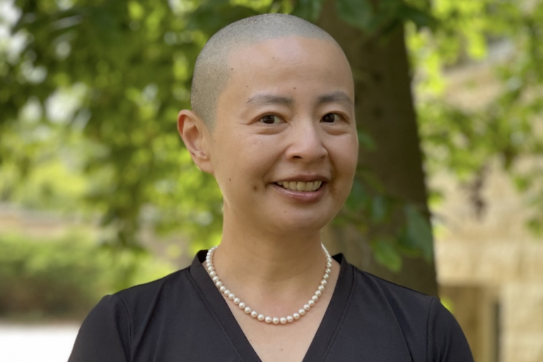 Person with a shaved head wearing a black v-neck shirt and pearl necklace, smiling in front of a background of green trees.