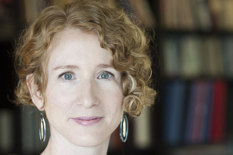 Close-up portrait of a person with curly light brown hair and large oval-shaped earrings, with a bookshelf blurred in the background.