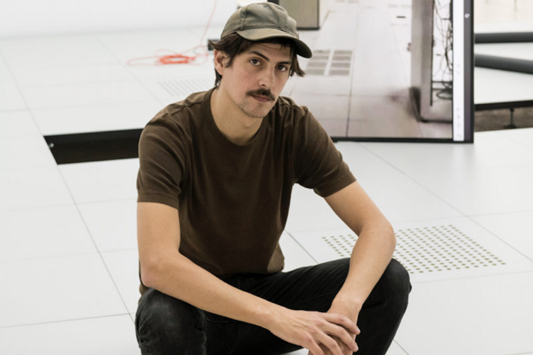 Man crouching indoors with a tiled floor background, a vertical monitor, and an extension cord.