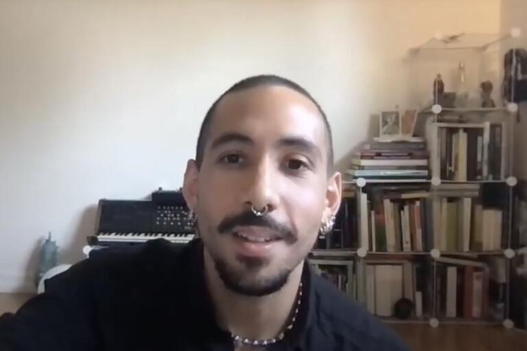 Person with shaved head and piercings, seated indoors with keyboard and bookshelves in the background.