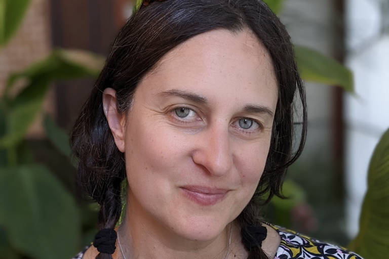 A woman with braided dark hair and blue eyes smiling in front of a blurred background with green plants.