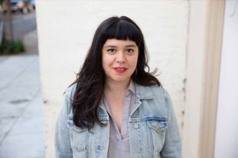 Woman with long dark hair and bangs wearing a light blue denim jacket over a gray top stands outdoors against a white wall.