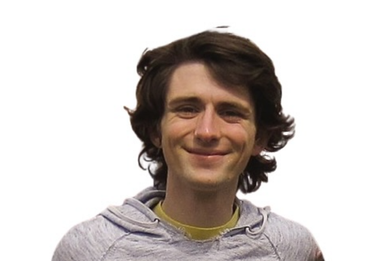Person with wavy brown hair wearing a light gray hoodie over a yellow shirt against a white background.
