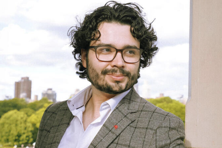 Man with glasses wearing a suit jacket and collared shirt, smiling outdoors with green foliage background.