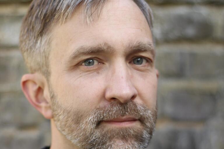 Close-up portrait of a middle-aged person with short light brown and grey hair, blue eyes, and a trimmed beard. Background is an out-of-focus stone wall.