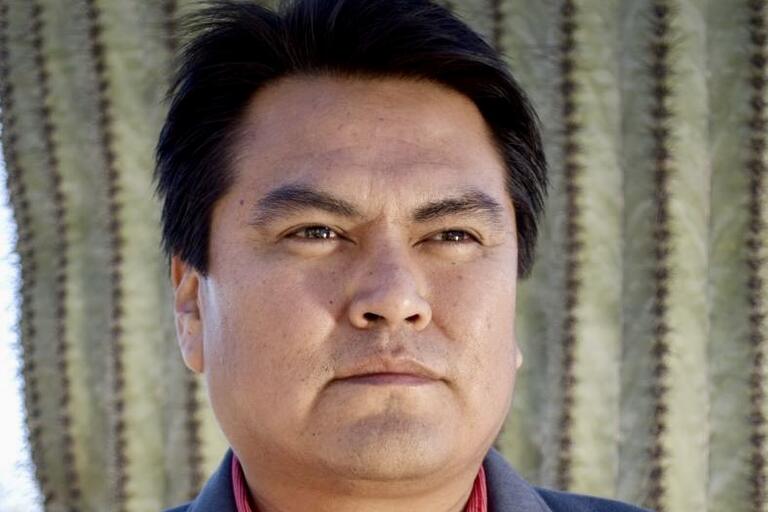 Close-up of a man's face with a serious expression in front of a blurred cactus background.