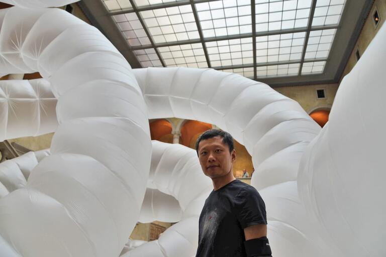 Man standing in front of large white inflatable tubular structures in an indoor gallery with high windows.